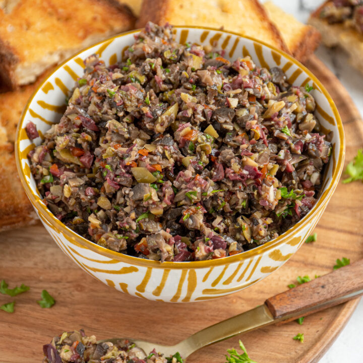 A wooden platter with toasts and a yellow and white bowl filled with homemade olive tapenade.