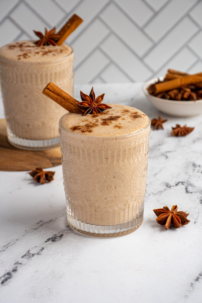 Glass cup filled with light brown banana chai smoothie garnished with cinnamon stick, star anise, and a dusting of spices.