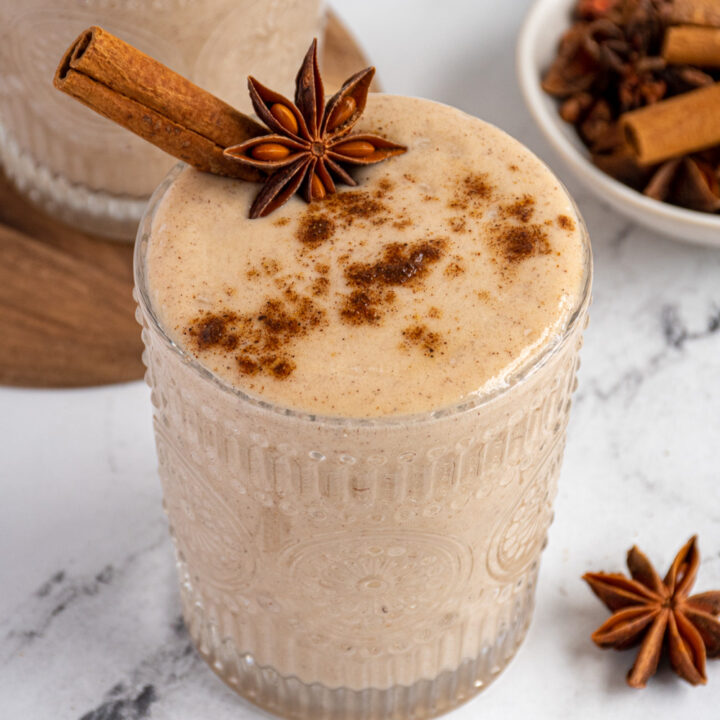 Close up of a light brown banana chai spice smoothie in a glass garnished with a cinnamon stick, star anise, and a dusting of ground spices.