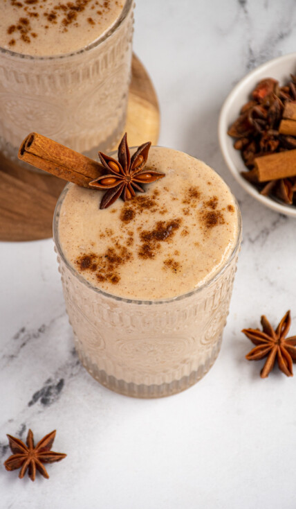 A glass filled with light brown banana chai smoothie garnished with a cinnamon stick, star anise, and a dusting of ground spices.