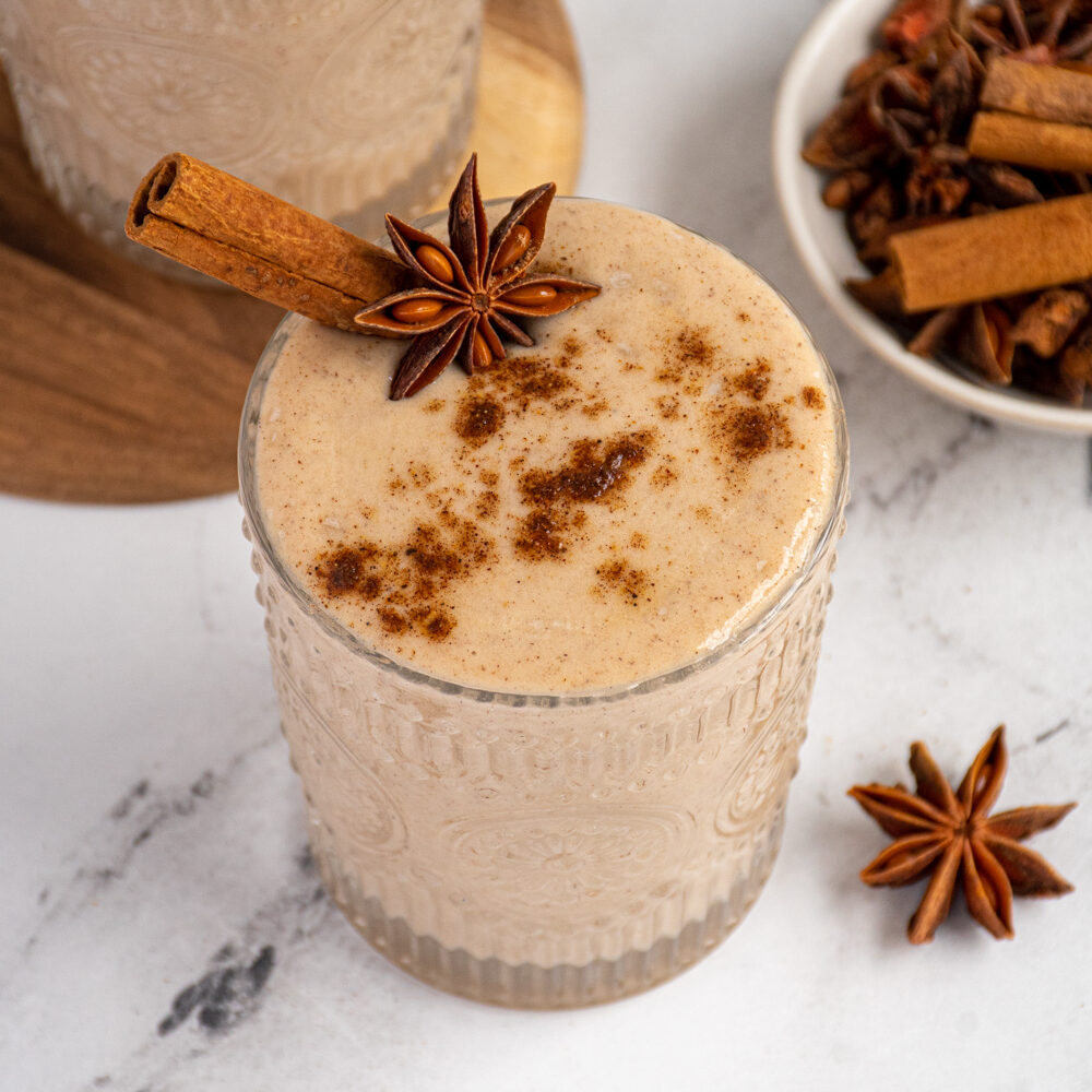A glass filled with light brown banana chai smoothie garnished with a cinnamon stick, star anise, and a dusting of ground spices.