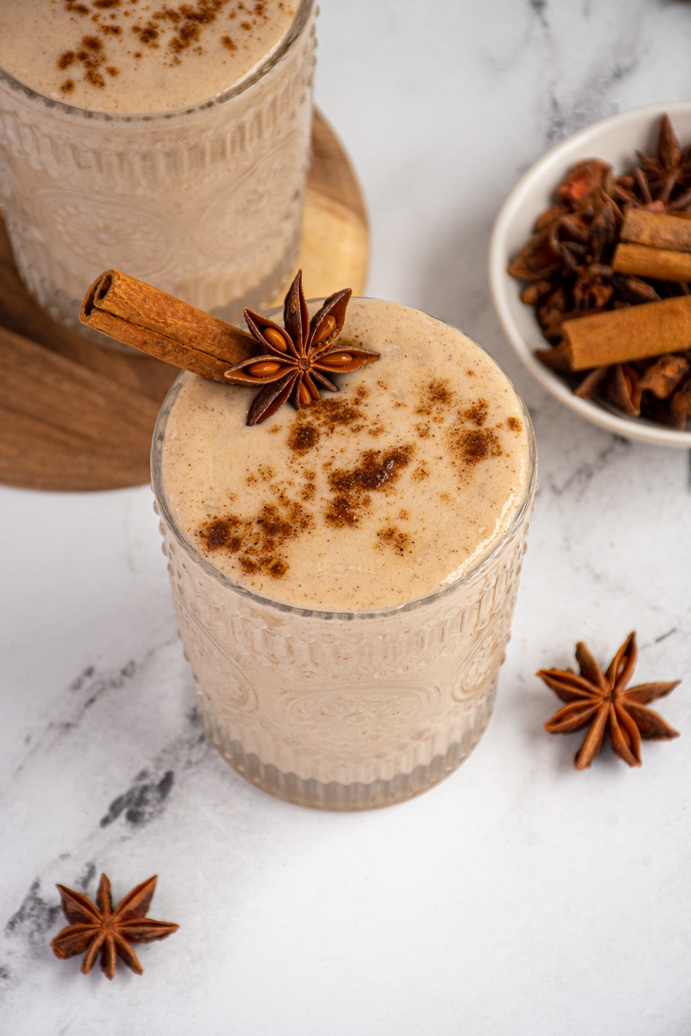 A glass filled with light brown banana chai smoothie garnished with a cinnamon stick, star anise, and a dusting of ground spices.