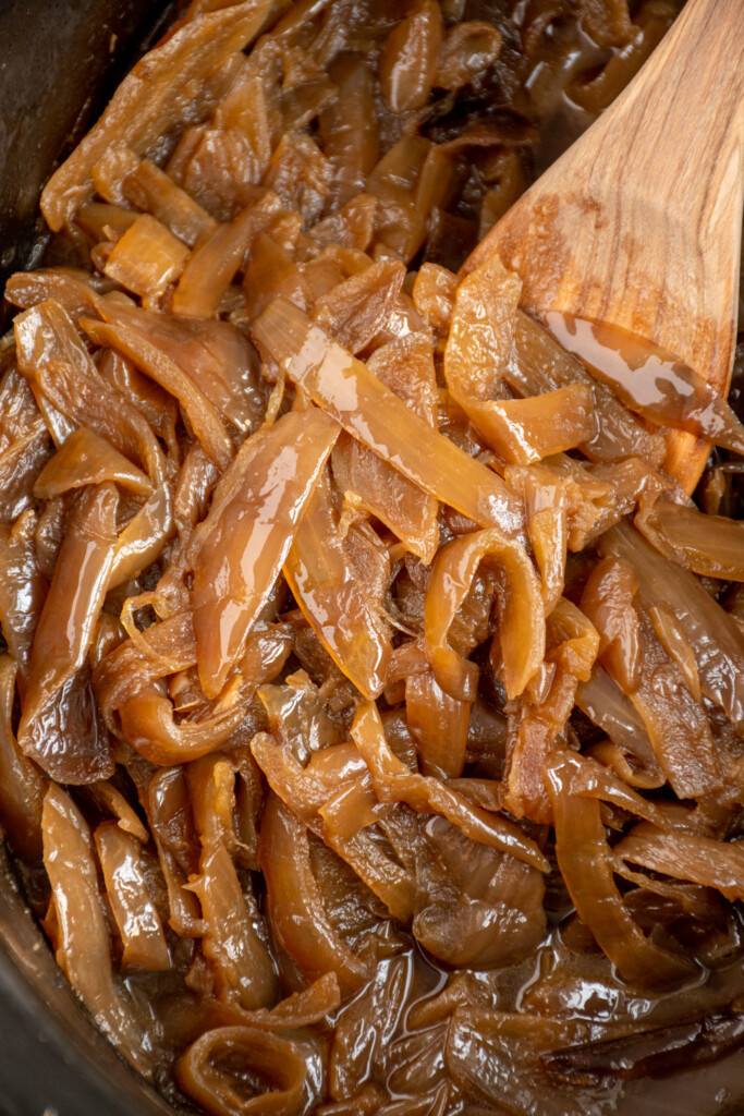 Close up of golden brown onions in a black crockpot with a wooden spoon.