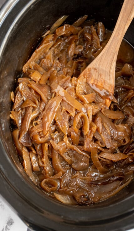 A black crockpot with slow cooker caramelized onions that a golden brown with a wooden spoon.