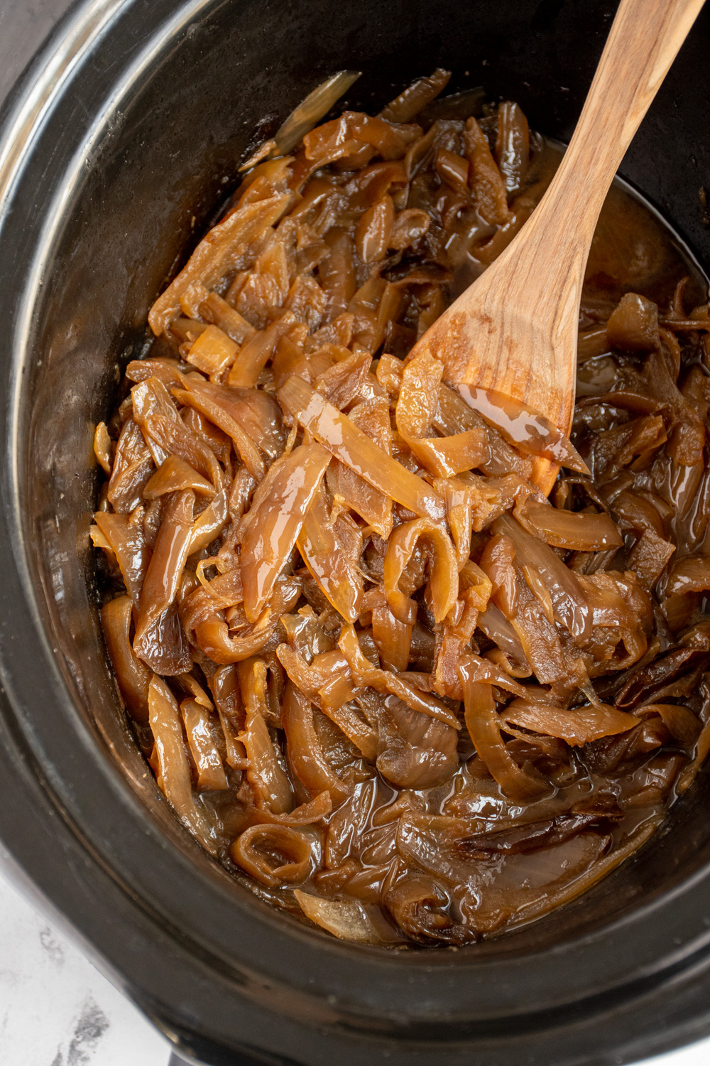 A black crockpot with slow cooker caramelized onions that a golden brown with a wooden spoon.