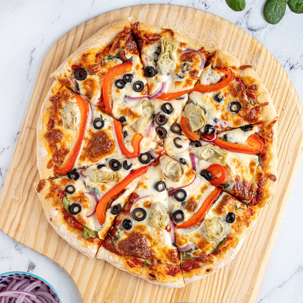 A homemade veggie pizza cut into pieces on a wooden pizza peel surrounded by pizza toppings.
