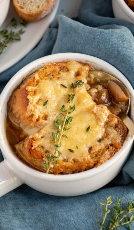 A white bowl filled with crockpot French onion soup and topped with baguette and cheese, garnished with thyme for serving.