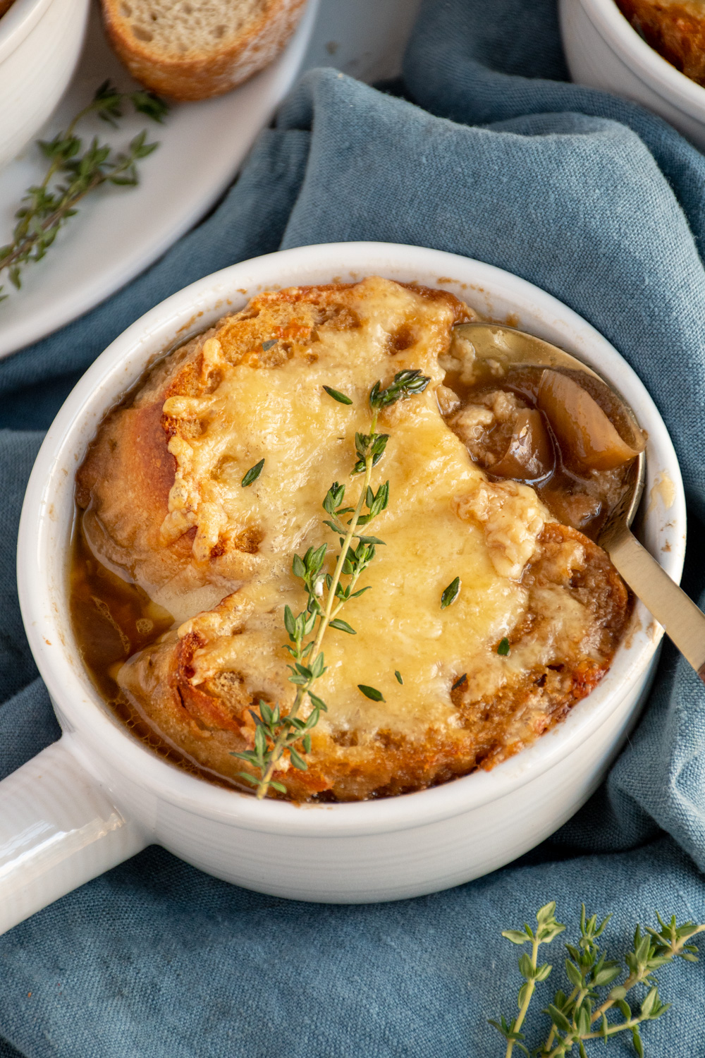 A white bowl filled with crockpot French onion soup and topped with baguette and cheese, garnished with thyme for serving.
