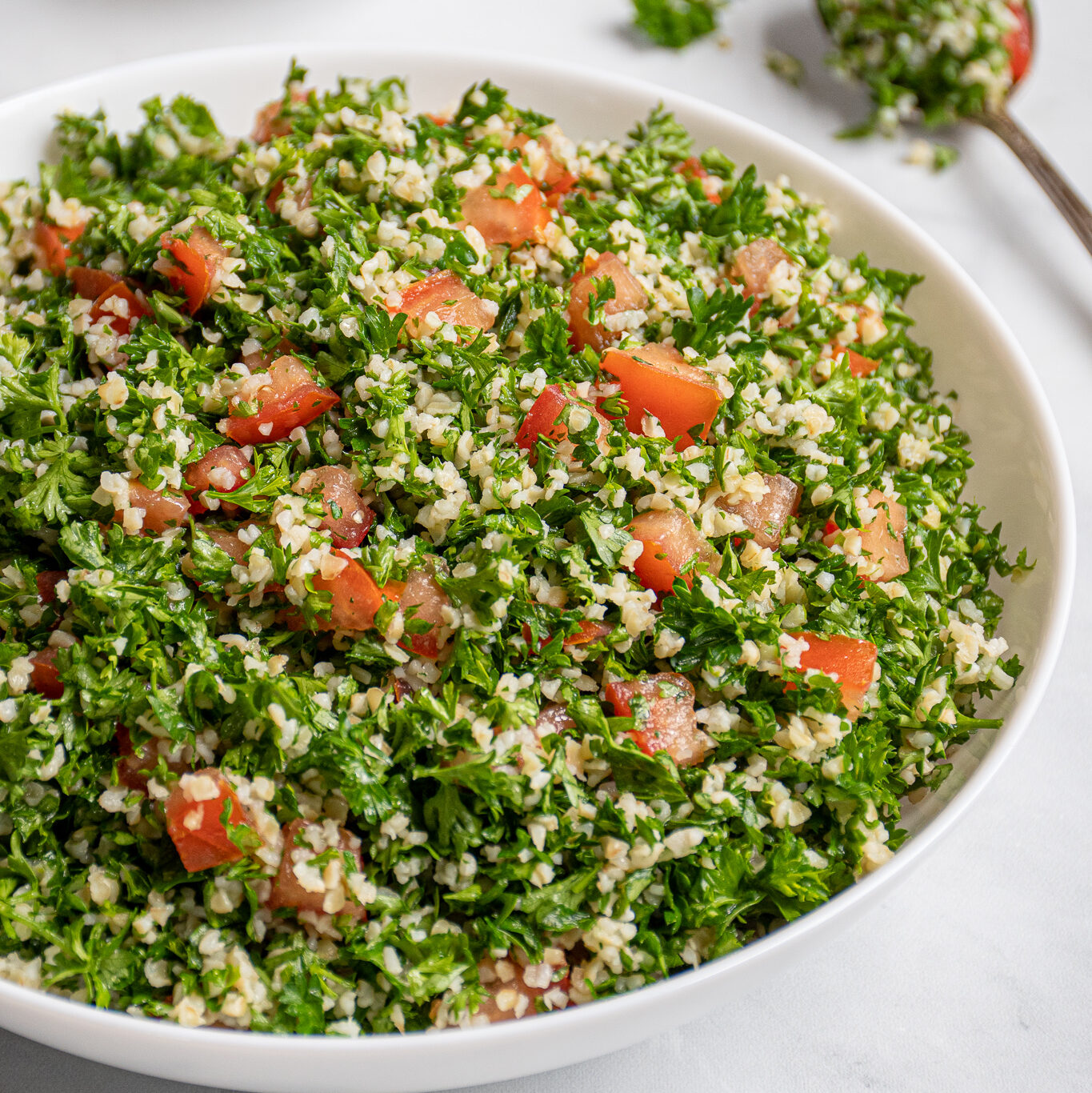 A large white bowl filled with tabbouleh salad with a spoonful out on the side with parsley leaves falling off.
