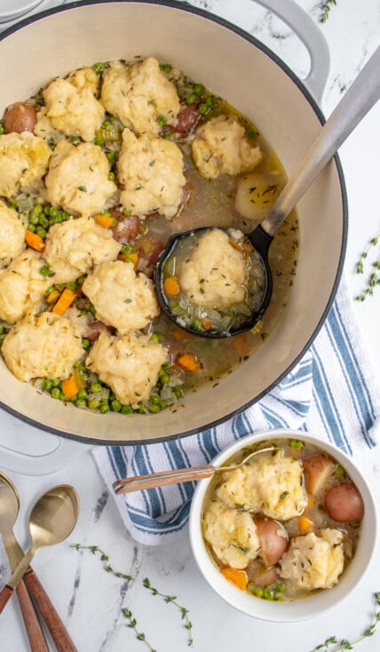 A dutch oven and bowl filled with veggie dumpling soup filled with carrots, celery, potatoes, and peas with fluffy dumplings on top.