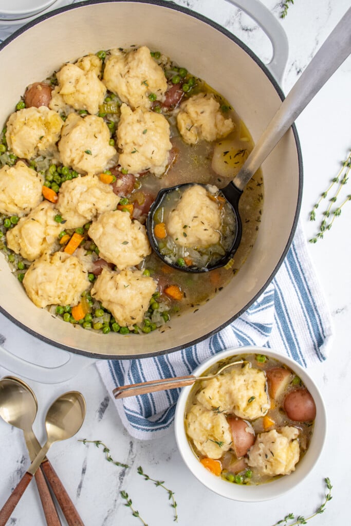 A dutch oven and bowl filled with veggie dumpling soup filled with carrots, celery, potatoes, and peas with fluffy dumplings on top.