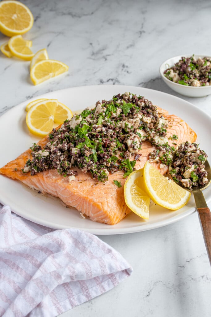 A plate with salmon with olive tapenade on top next to some lemon wedges.