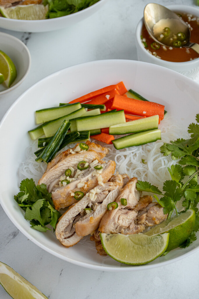A white bowl filled with white vermicelli noddles with lemongrass chicken and veggie to garnish.