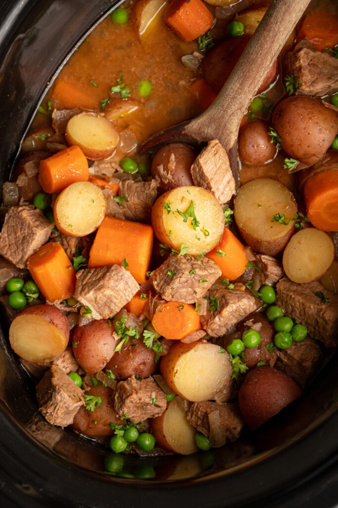 Close up of vegetables and stew meat in the crockpot with a wooden spoon scooping the beef stew.