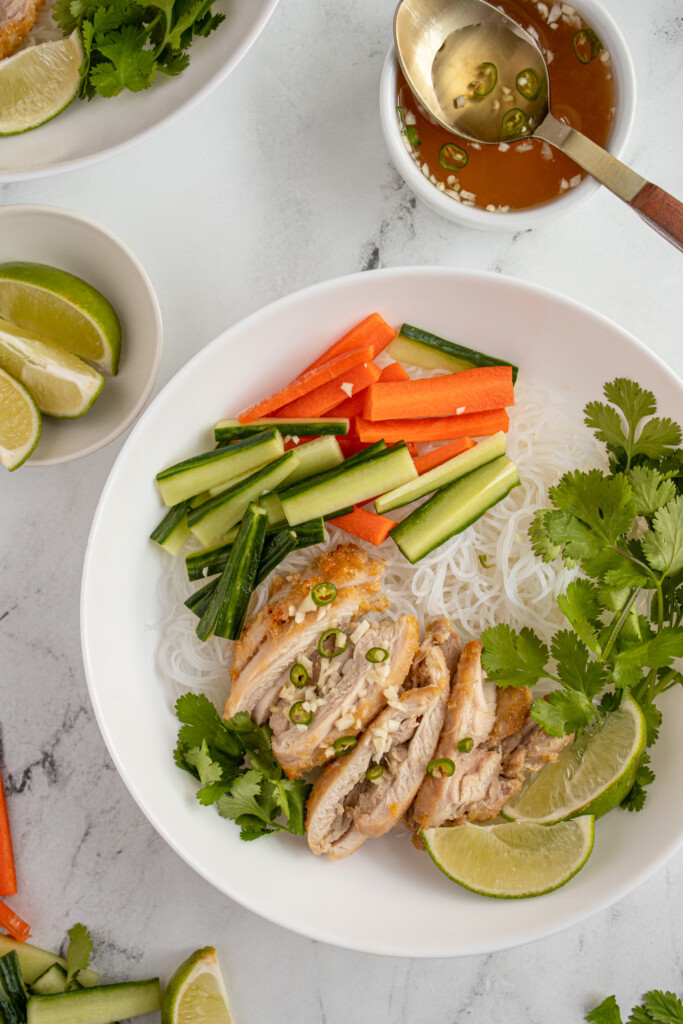 Overhead view of a large white bowl filled with vermicelli noodles and topped with lemongrass chicken, vegetables, and nuoc cham. 