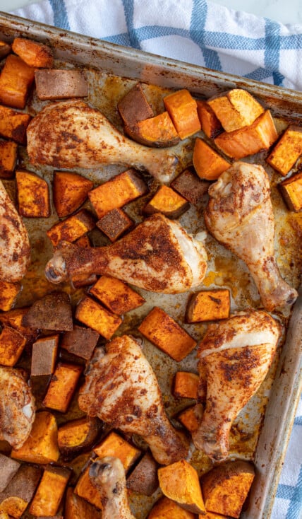 A sheet pan with baked chicken legs and sweet potato chunks covered in seasoning.