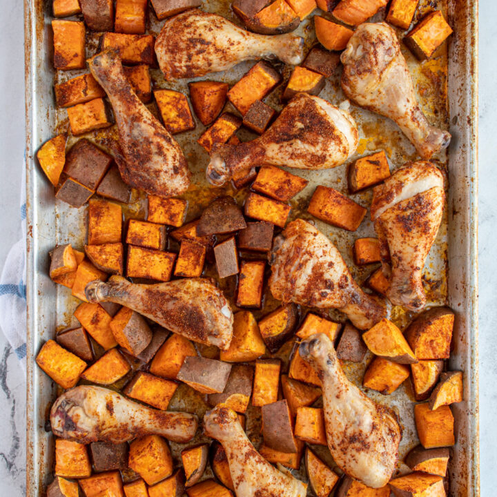 Overhead view of a sheet pan with baked chicken legs and sweet potatoes all covered in seasoning.
