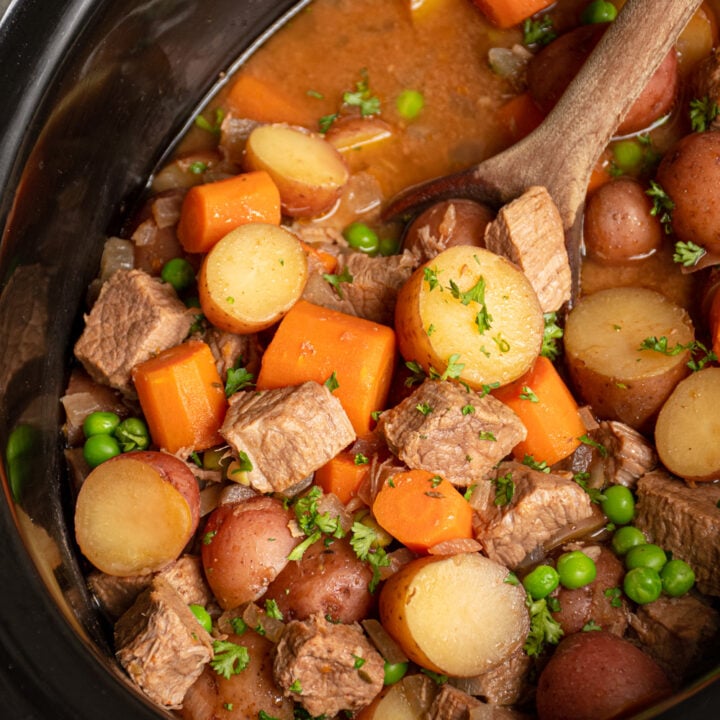 Black crockpot filled with beef stew with big chunks of vegetables and stew meat and a wood spoon scooping them.