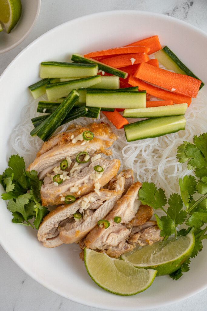 Overhead photo of a white bowl with lemongrass chicken not top of vermicelli noodles and vegetables with nuoc Cham sauce.