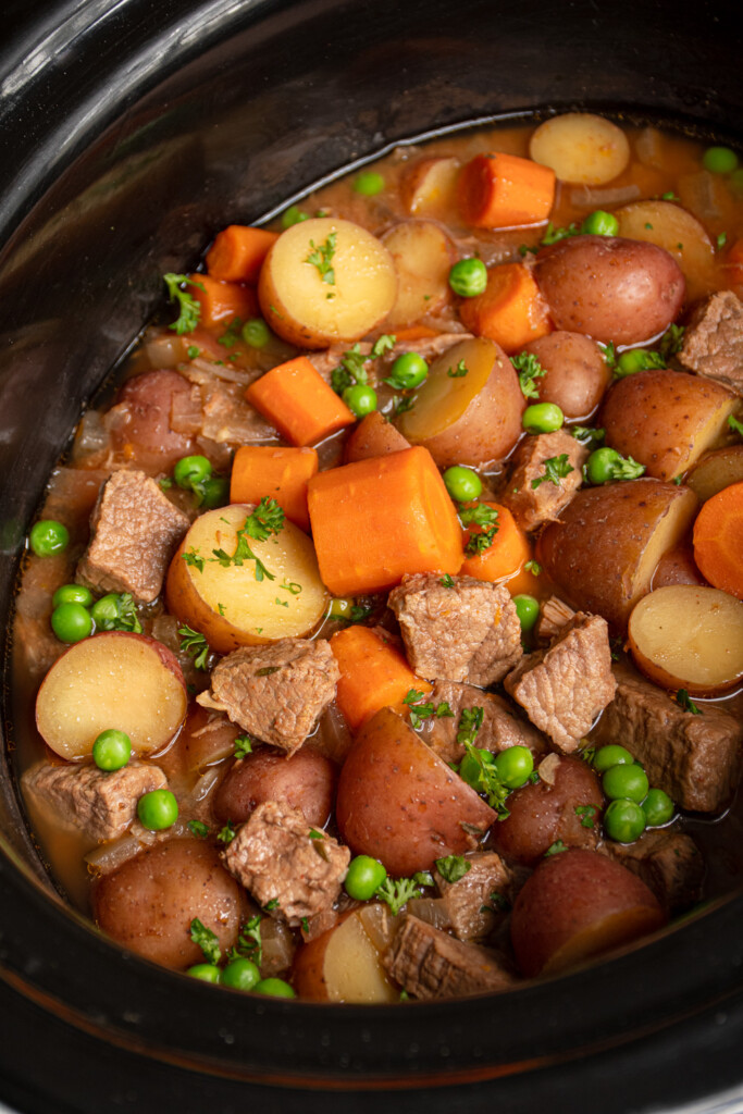 A slow cooker filled with beef stew with carrots, potatoes, peas, and beef.