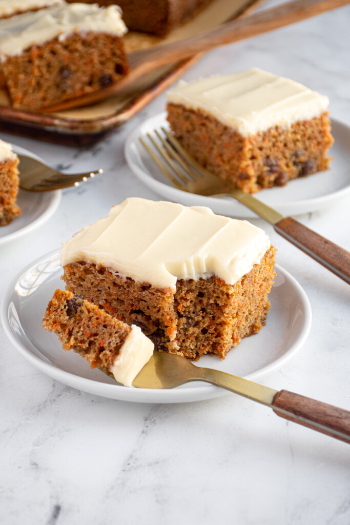 A white plate with a square slice of carrot cake with a bite on a fork taken out.