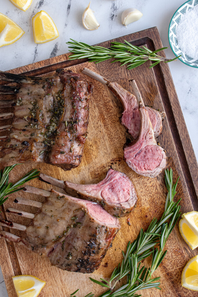 Wooden cutting board with two rack of grilled lamb with the first three pieces cut off showing a pink lamb interior.