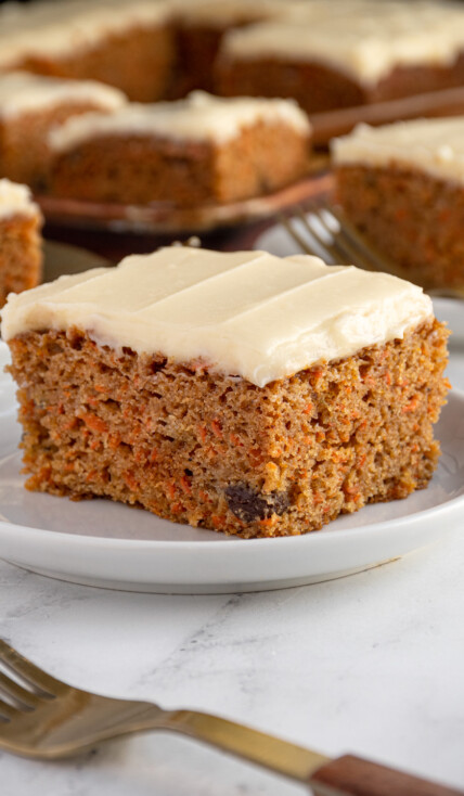 A white plate with a square cut piece of orange spiced easy carrot cake with orange flecks and raisins, topped with a cream cheese frosting.