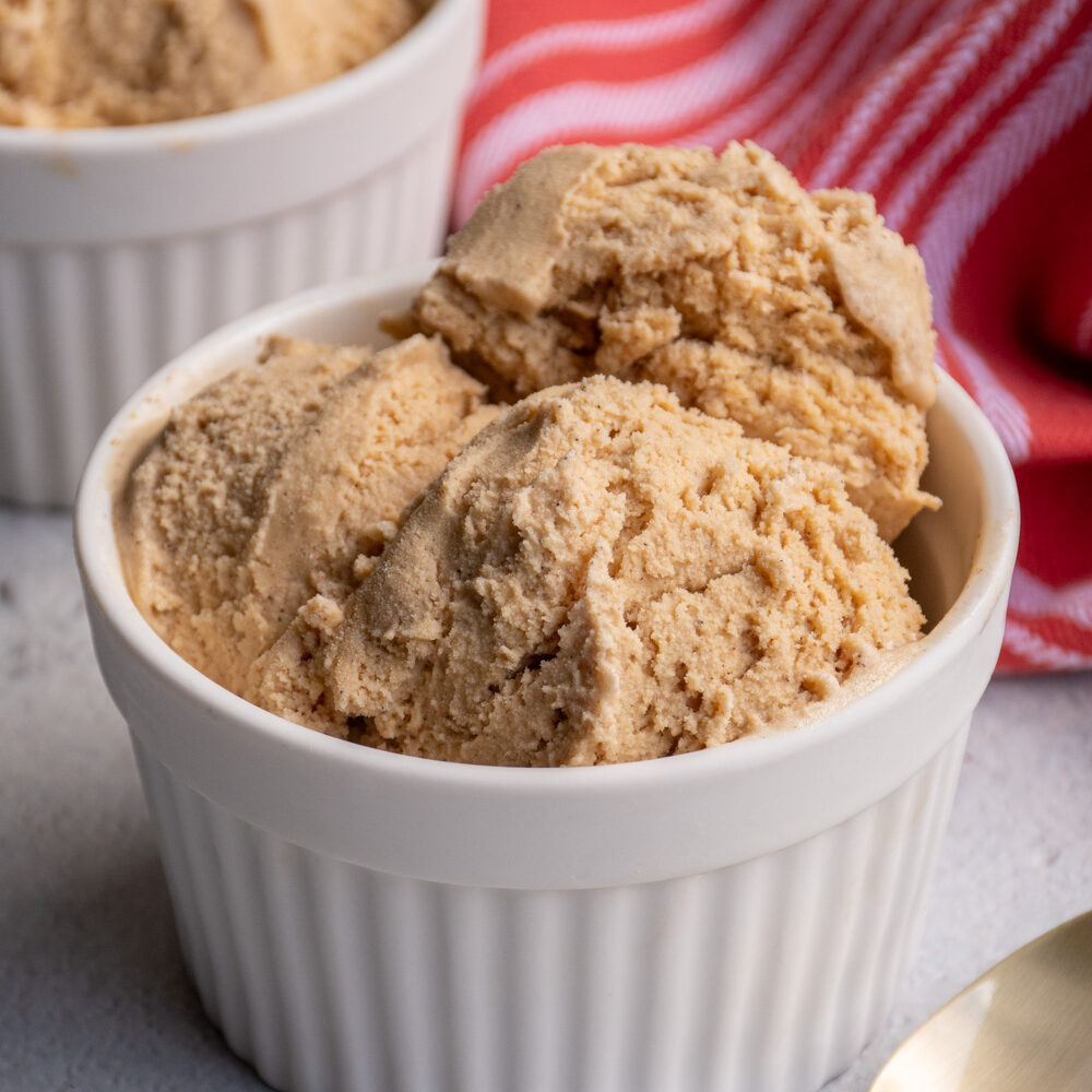 A white bowl filled with scoops of light brown colored gingerbread ice cream sitting next to a spoon.