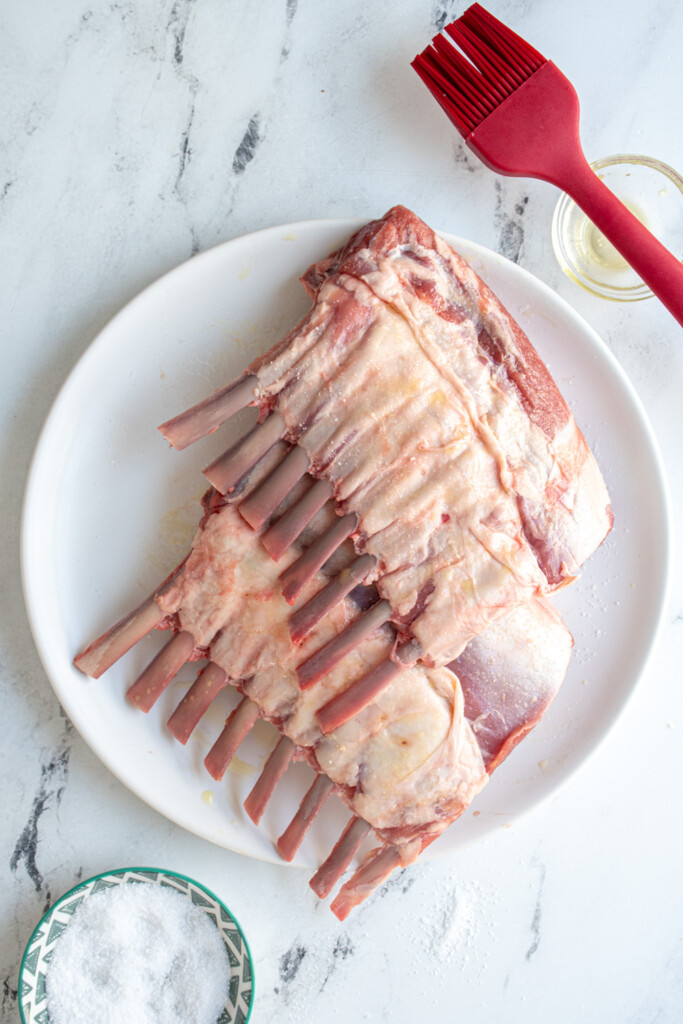 Oiled and salted rack of lamb of a white plate ready to be put on the grill.