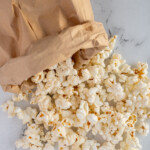 Pinterest pin with text overlay "Paper Bag Microwave Popcorn", image of a brown bag rolled up and popcorn tumbling out onto a tabletop.