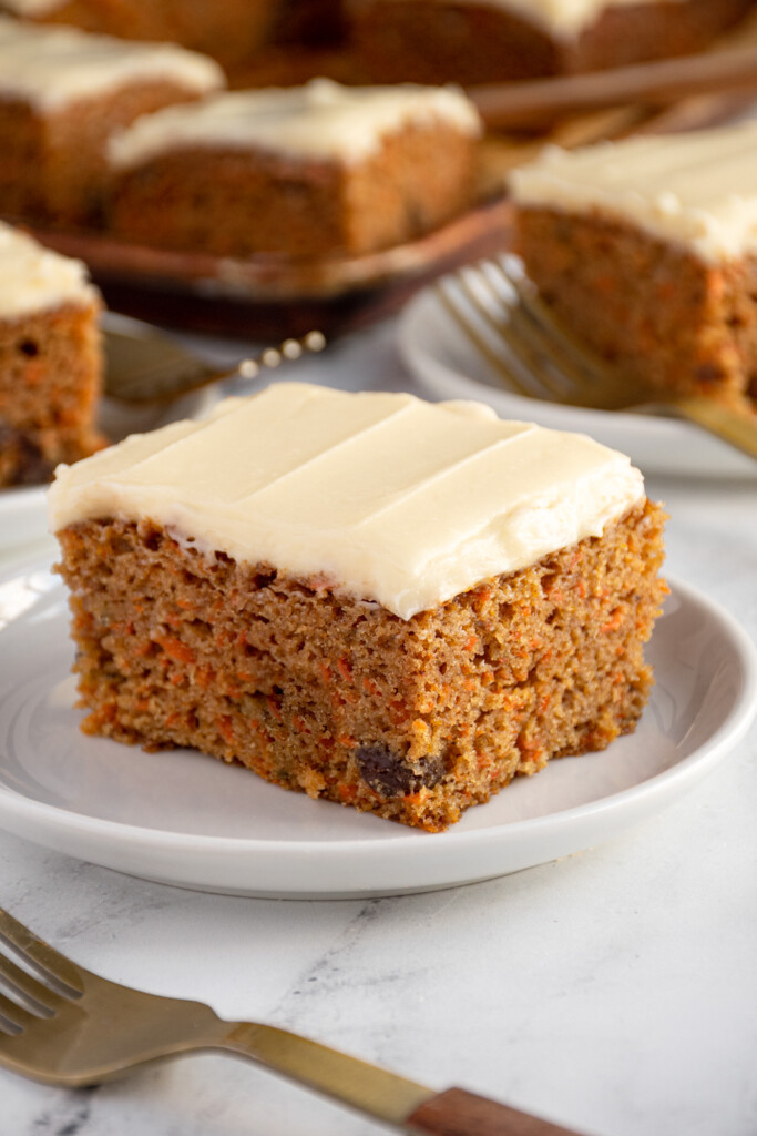 A white plate with a slice of square carrot cake with flecks of carrots and raisins topped with a cream cheese frosting.