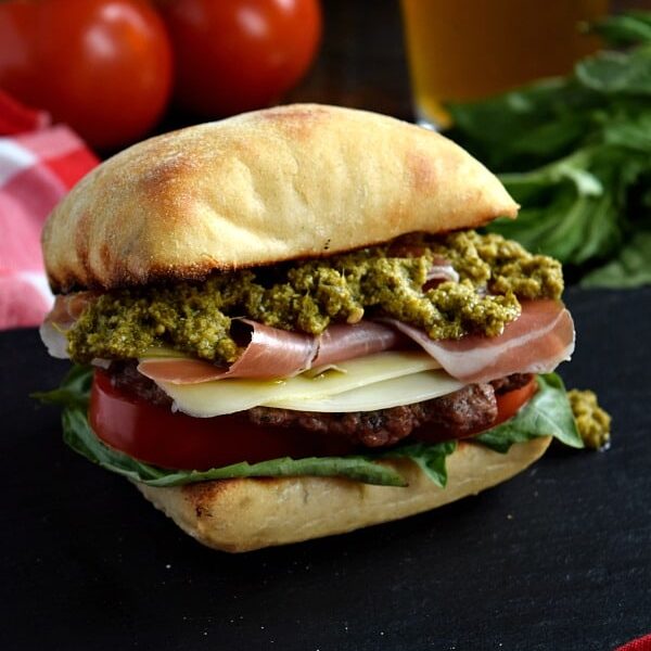 a close up of a prosciutto pesto burger on a black plate with a beer