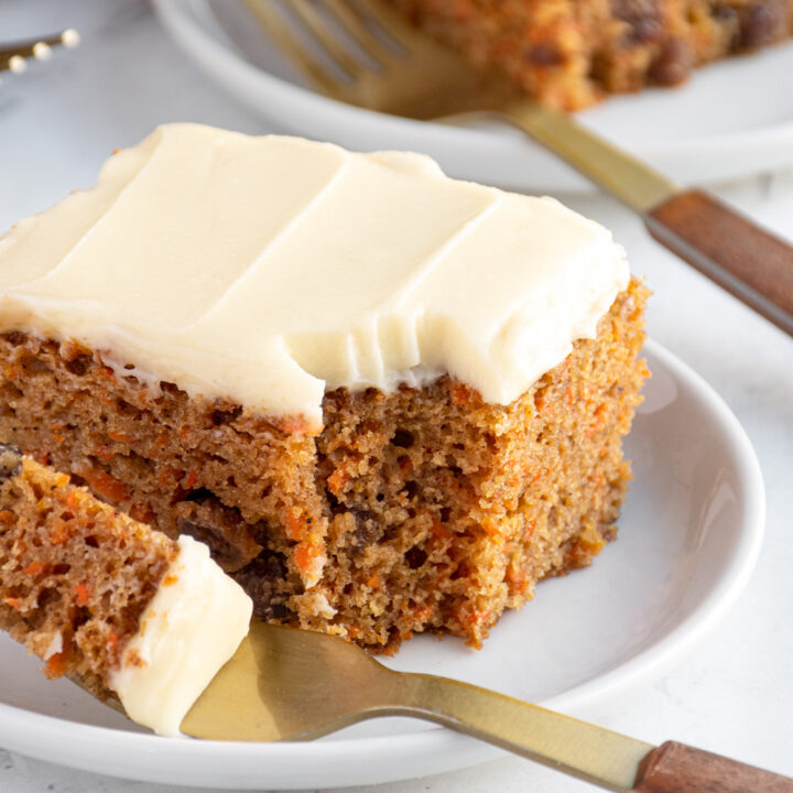 A close up of a slice of carrot cake on a white plate with a bite taken out on a fork sitting on the edge of the plate.