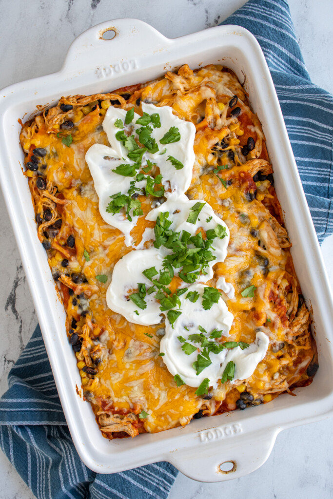 Overhead view of a white casserole pan filled with layered chicken enchilada casserole topped with sour cream and cilantro.