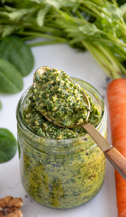 A glass jar with a spoon resting on top filled with bright green carrot top pesto.