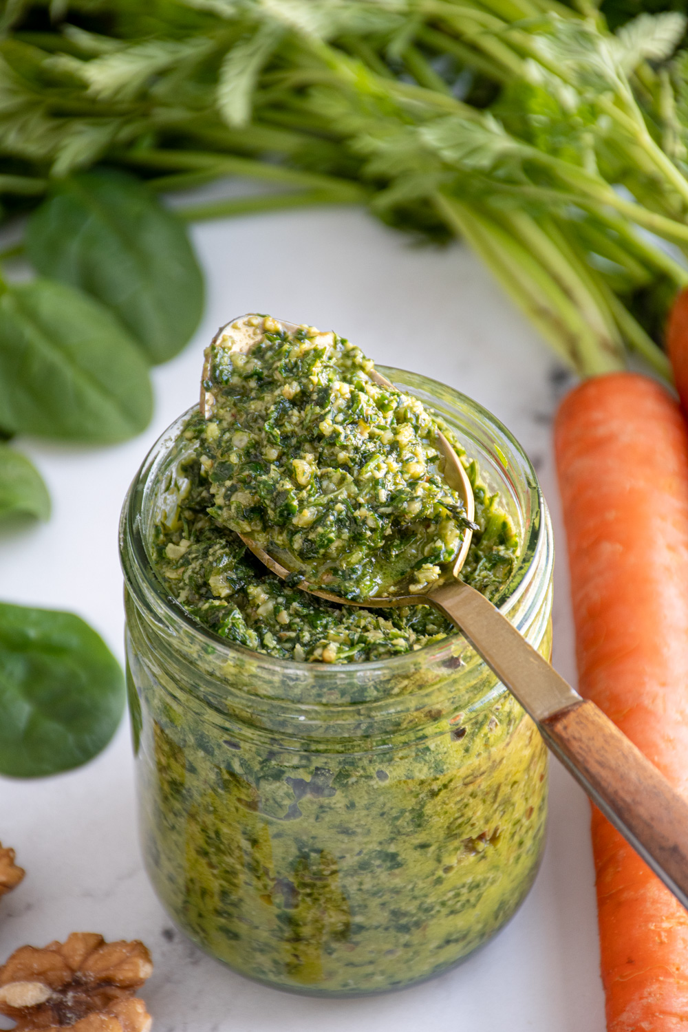 A glass jar with a spoon resting on top filled with bright green carrot top pesto.