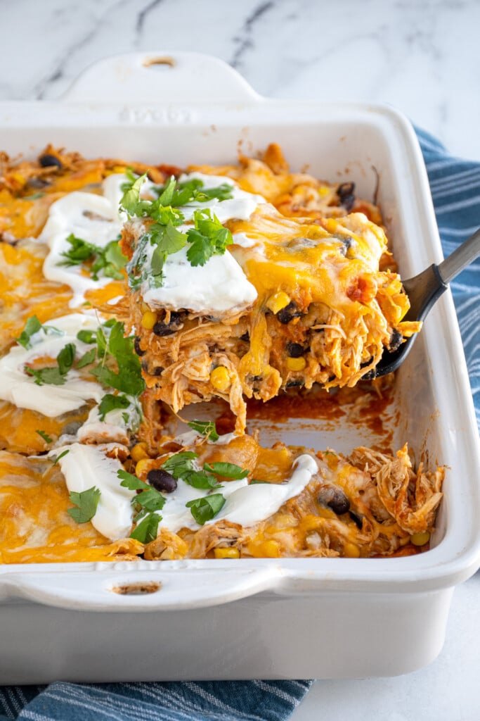 A spatula with a large piece of chicken enchilada casserole on it over a baking dish showing off chicken, black beans, and corn.