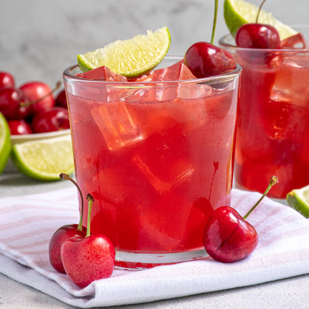 A glass filled with ice and bright red cherry margarita topped with a fresh cherry and lime wedge.
