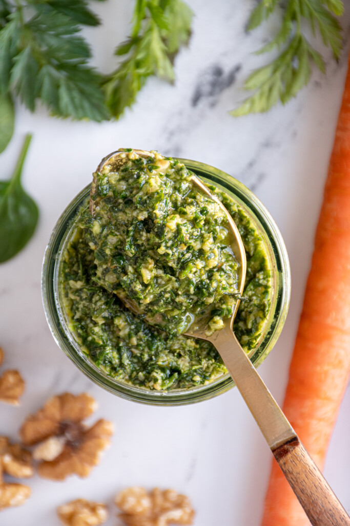 overhead look at a glass jar with a spoon resting on the top filled with carrot top pesto.