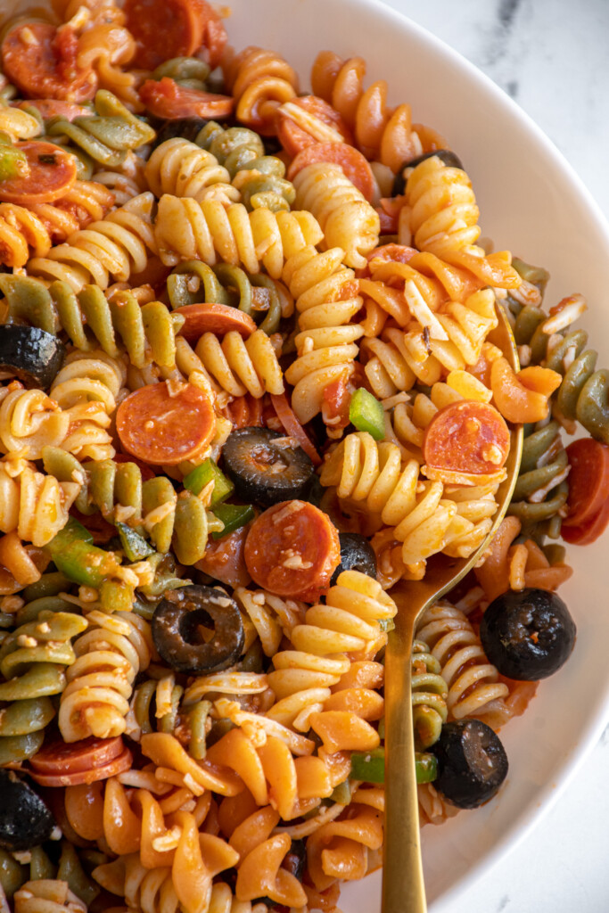 Close up of pizza pasta salad in a white bowl with a gold spoon.