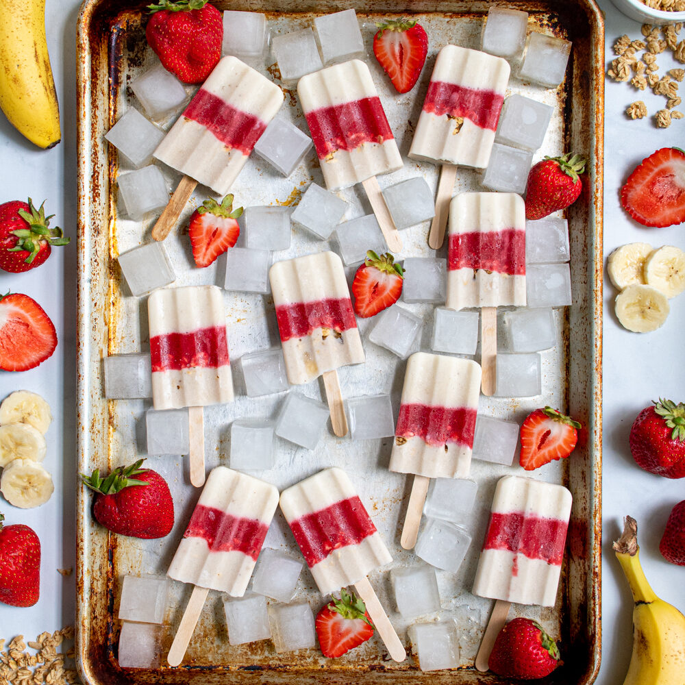 Sheet tray full of red and white parfait popsicles and ice cubes surrounded bu sliced strawberries, bananas, and bits of granola.