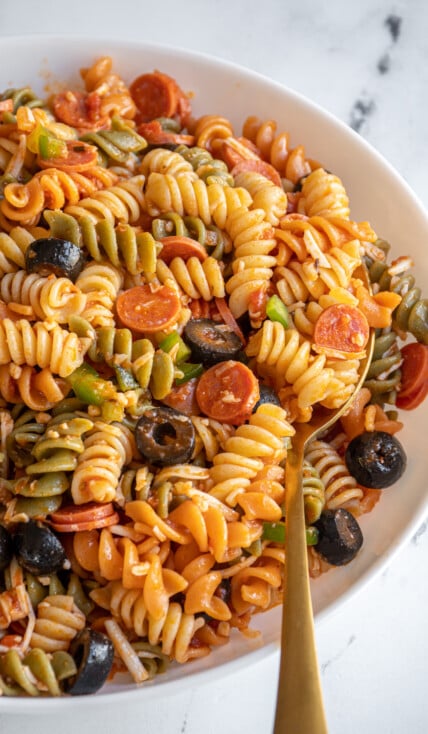 A white bowl with a gold spoon in pasta pizza salad.