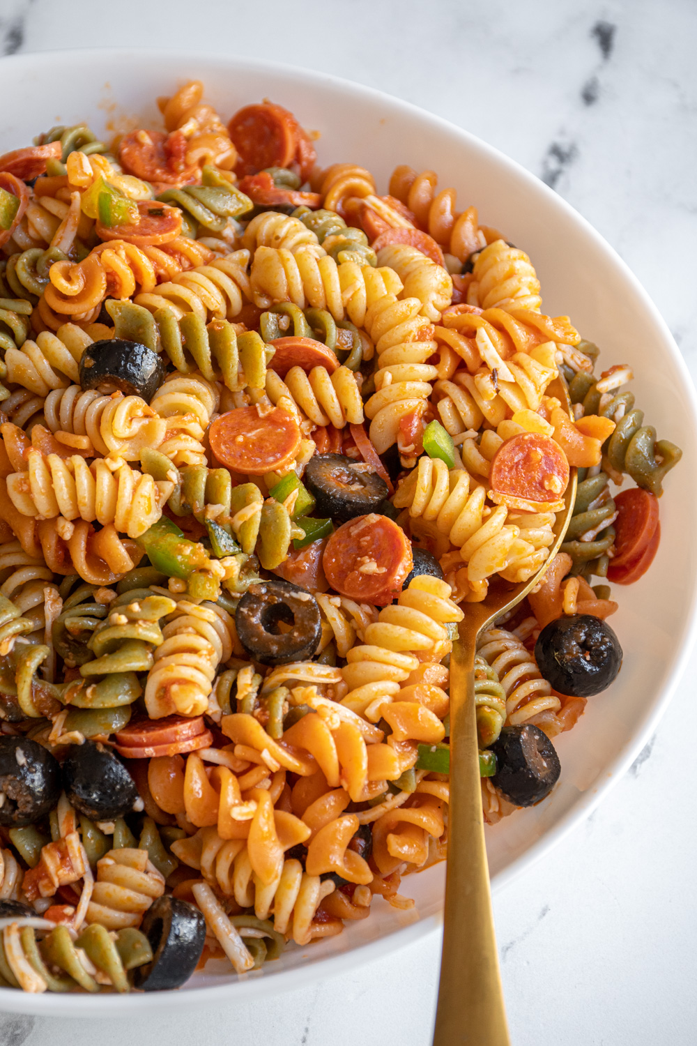 A white bowl with a gold spoon in pasta pizza salad.