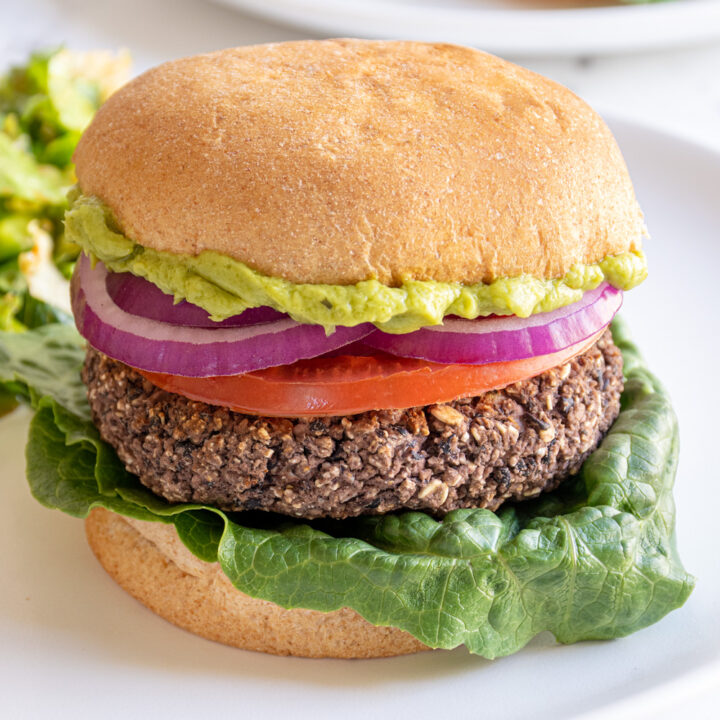 A plate with a grilled black bean burger in a bun with lettuce, tomato, red onion, and guacamole.