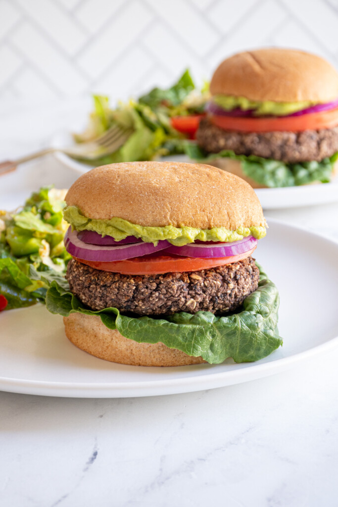 A white plate with a grilled black bean burger in a bun with lettuce, tomato, red onion, and guacamole.
