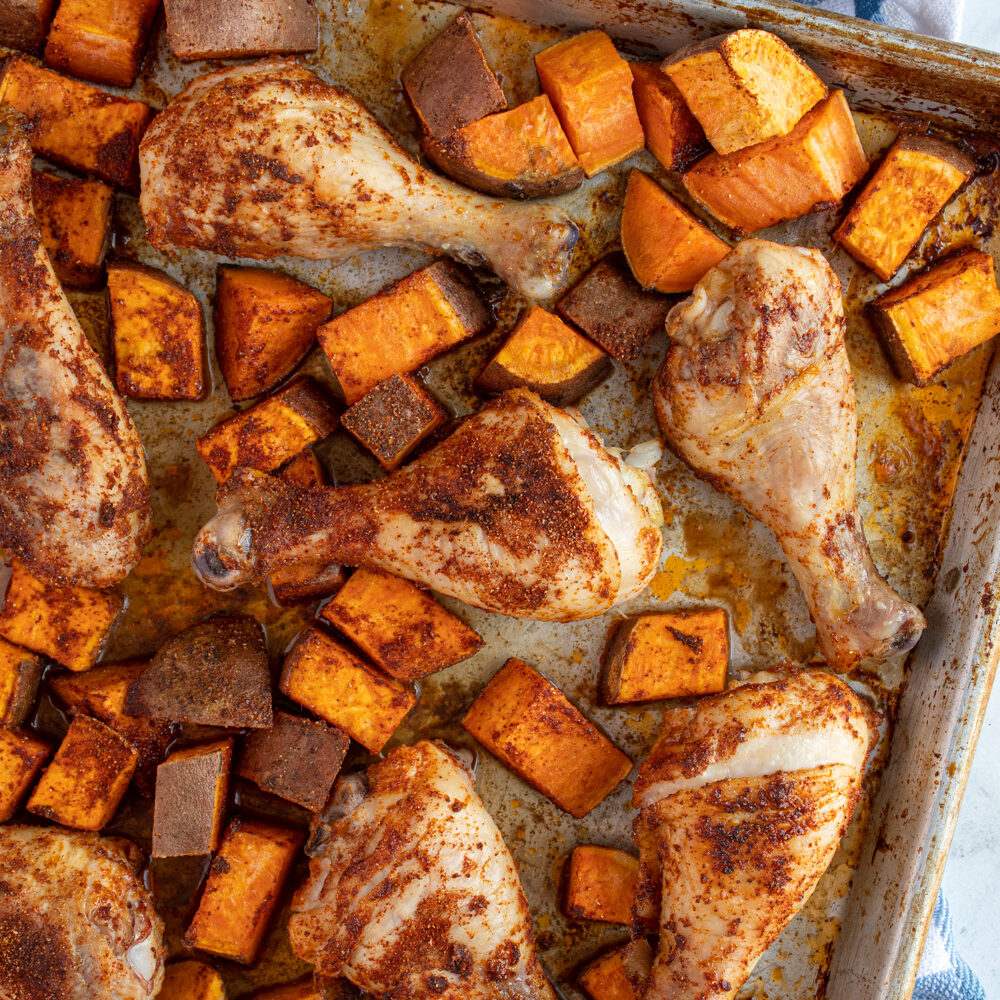 A sheet pan with baked chicken legs and sweet potato chunks covered in seasoning.