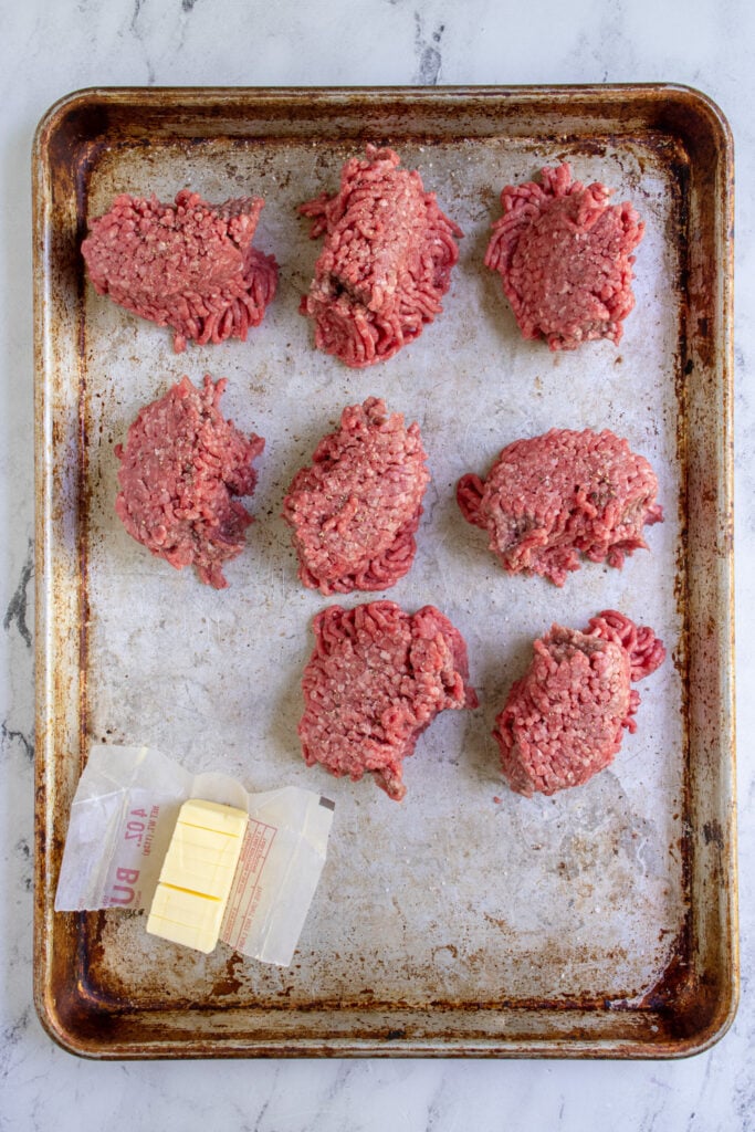 Eight pieces of ground beef and butter cut into pieces on a sheet pan all prepped for being cooked on the grill.
