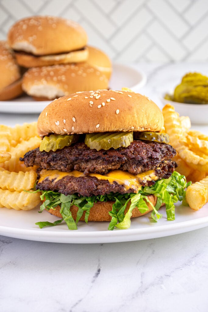 A white plate with French fries and a double smash burger in a bun with lettuce and pickles.