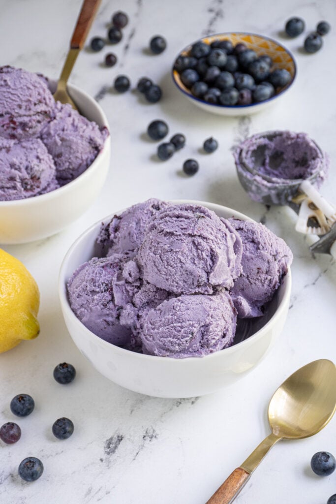 A bowl of homemade blueberry ice cream next to a spoon and ice cream scoop.