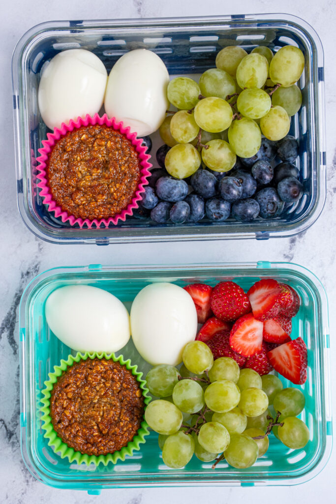 Two container of burst boxes sitting on a table with eggs, muffins, and fruit.